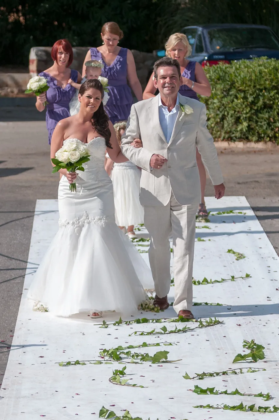 Bride in a white strapless gown holding a bouquet of white roses walking arm-in-arm with a man in a light-colored suit down an outdoor aisle decorated with green leaves.