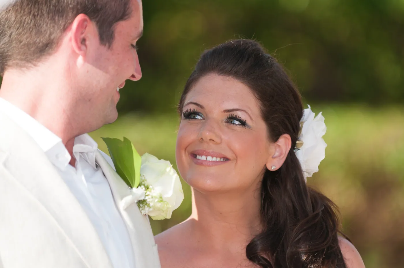 Bride with long dark hair and white flower looking lovingly at groom in light-colored suit with white rose boutonniere.