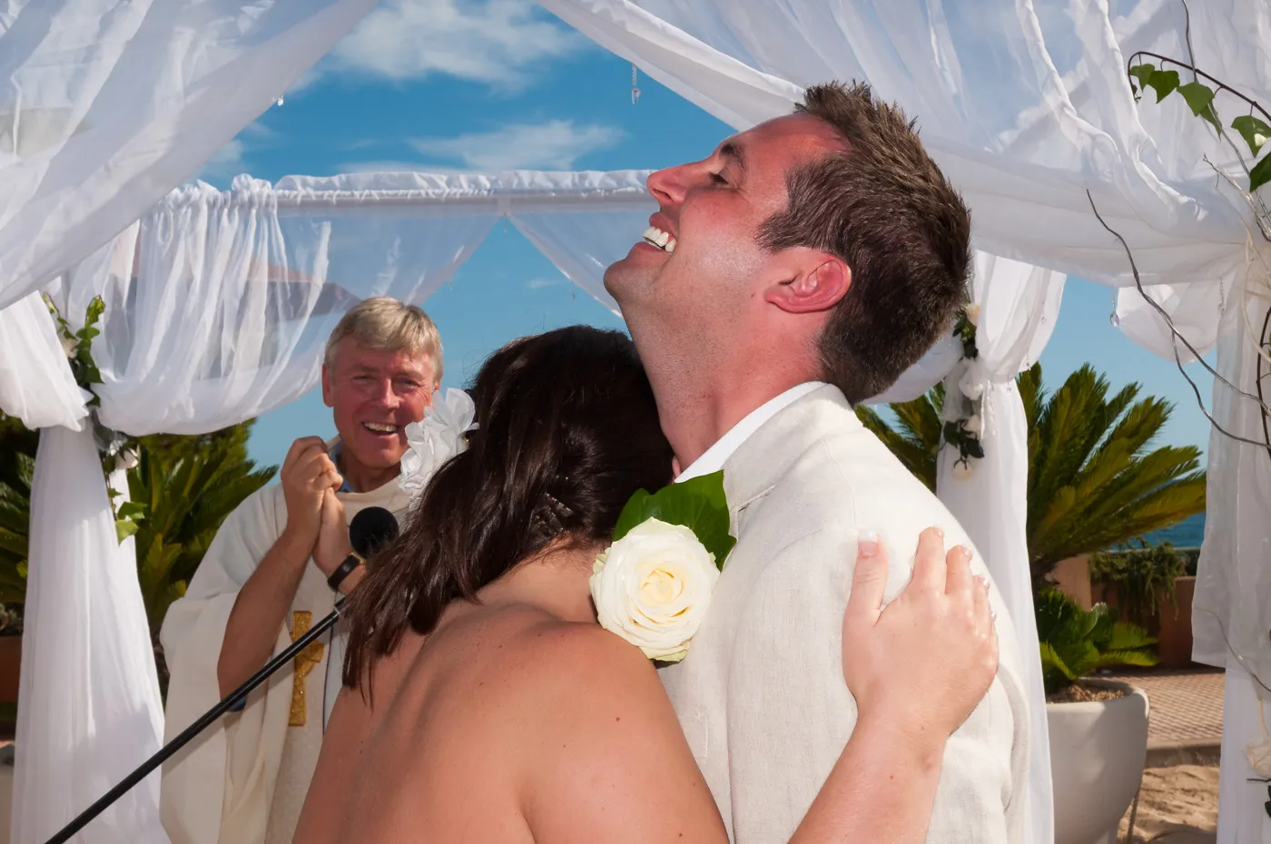 Newlywed couple embraces joyfully under a white canopy at an outdoor wedding ceremony with a smiling officiant in the background.