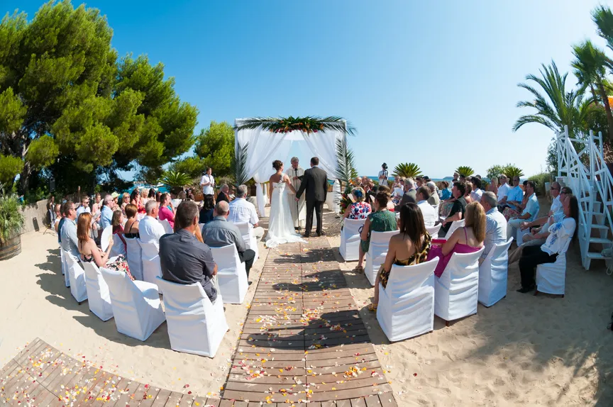 Outdoor beach wedding ceremony with bride and groom holding hands under a white floral arch, guests seated on covered chairs on sand.
