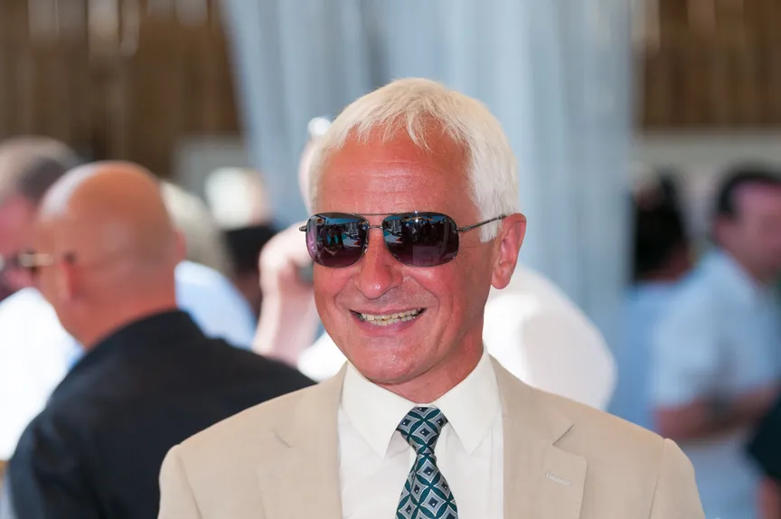 Smiling older man with white hair wearing sunglasses, a beige suit, white shirt, and patterned tie at a social event.
