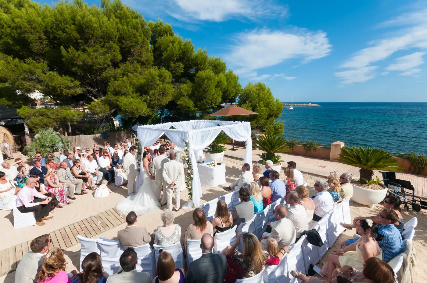 Outdoor beachside wedding ceremony with guests seated in white-covered chairs facing a bride and groom under a white canopy, with the sea and blue sky in the background.