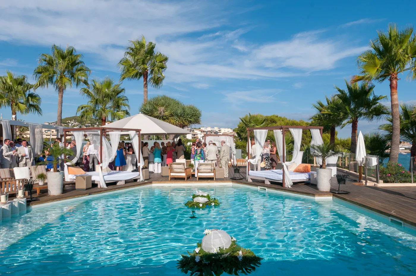 Outdoor pool area with cabanas and lounge chairs surrounded by palm trees and people gathered near the back under blue sky.