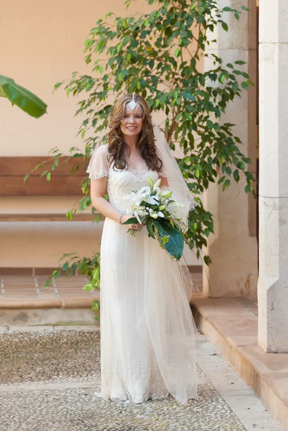Woman in a white wedding dress holding a bouquet of white flowers standing outdoors near a green plant and stone wall.