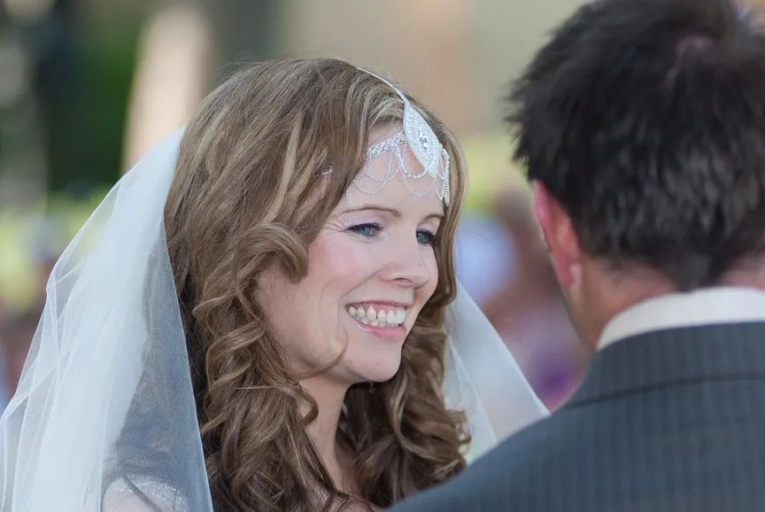 Bride smiling and looking at groom during wedding ceremony, wearing a white veil and decorative headpiece.