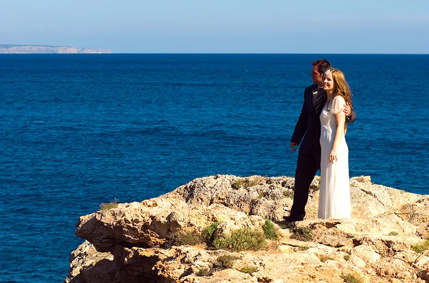Bride and groom standing on rocky cliff overlooking a deep blue sea during daytime.