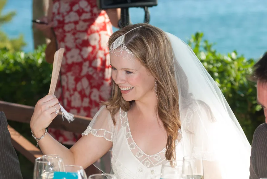 Smiling bride in a lace wedding dress and veil holding a handheld fan at an outdoor reception.