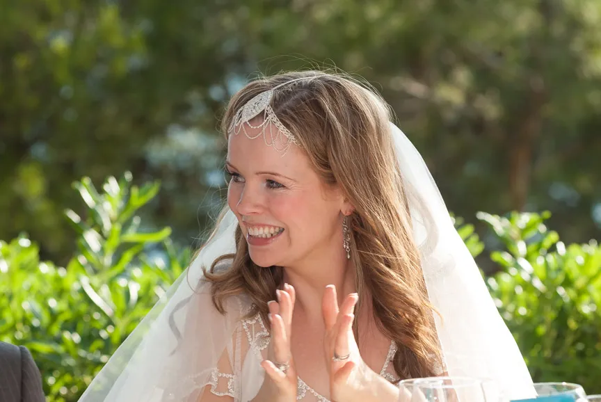 Smiling bride in a white wedding dress and veil clapping her hands outdoors with greenery in the background.