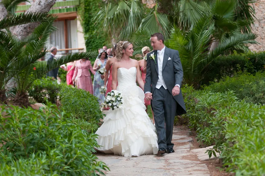 Bride and groom holding hands and walking on a stone path surrounded by lush greenery and wedding guests in the background.