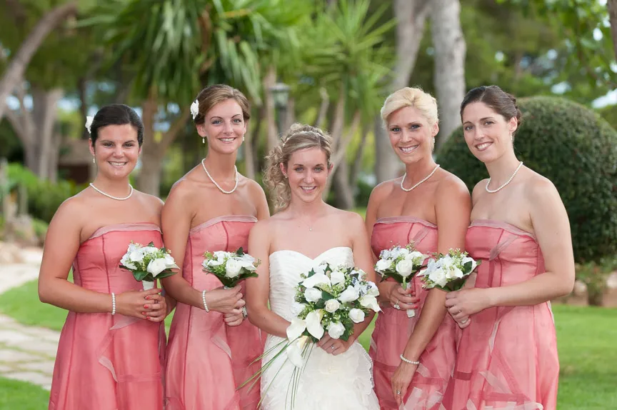 Bride in white wedding dress standing outdoors with four bridesmaids in matching pink strapless dresses holding white floral bouquets.