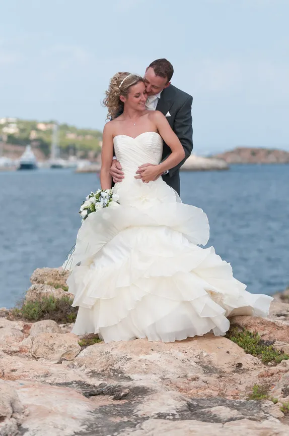 Bride and groom embracing on rocky shore with ocean and boats in the background.