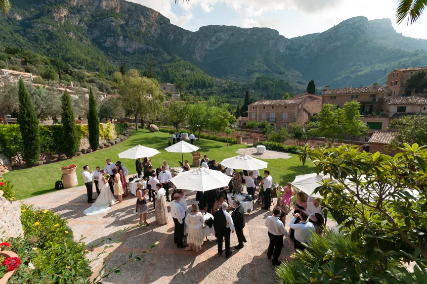 Outdoor wedding reception with guests gathered around tables under white umbrellas in a garden with mountains and rustic buildings in the background.