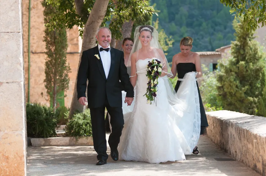 Bride in white wedding gown holding a cascading bouquet, walking hand in hand with a man in a black tuxedo, followed by two bridesmaids outdoors.