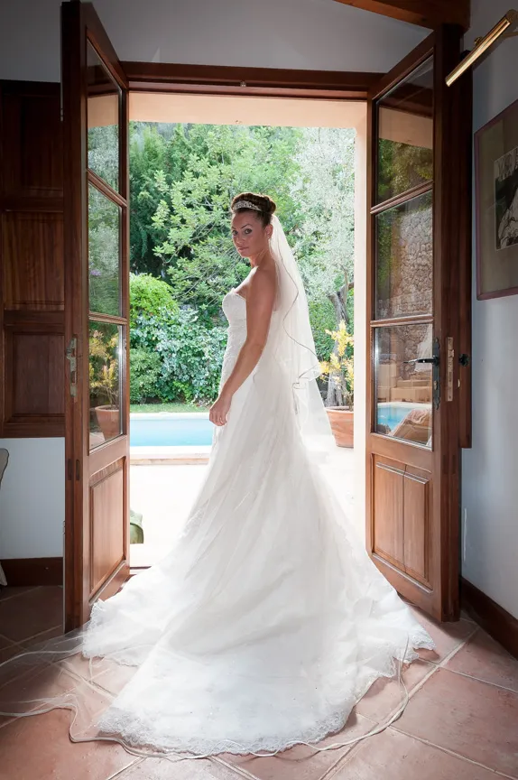 Bride in a white wedding gown and veil standing in an open doorway with greenery and a pool visible outside.