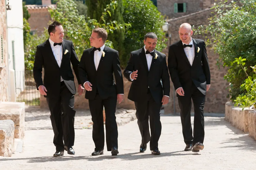 Four men in black tuxedos walking and talking on a sunny stone pathway with greenery and a rustic building in the background.