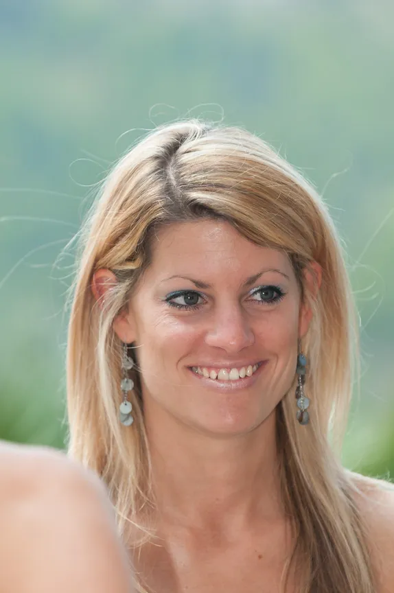 Smiling woman with long blonde hair wearing dangling earrings and natural makeup against a blurred outdoor background.