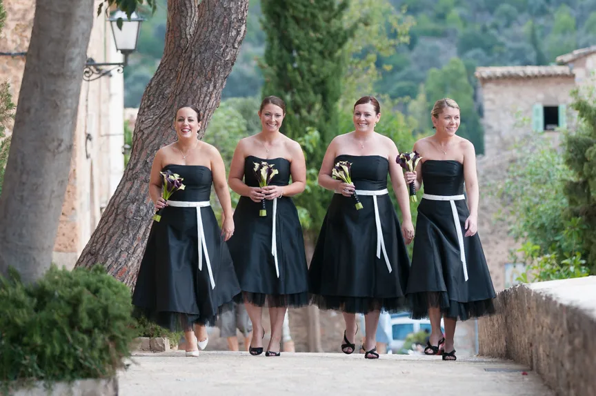 Four bridesmaids in strapless black dresses with white sashes holding small bouquets, walking outdoors on a stone path.