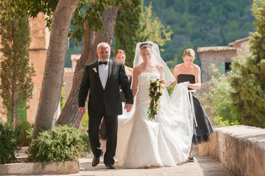Bride in a white wedding gown holding a cascading bouquet, walking outdoors with a man in a tuxedo and two bridesmaids assisting her dress.