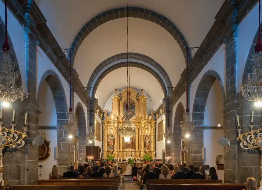 Interior of a stone-arched church with an ornate golden altar and a bride and groom seated with guests watching.