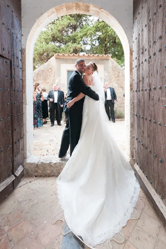 Bride and groom embracing and smiling under a stone archway with wooden doors, with guests in the background.
