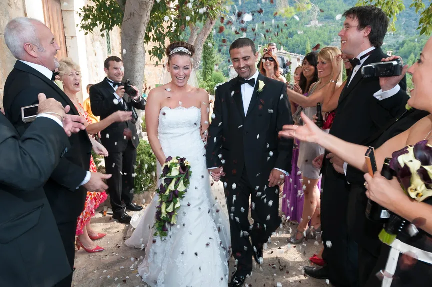 Bride and groom smiling and holding hands while guests throw flower petals during an outdoor wedding celebration.