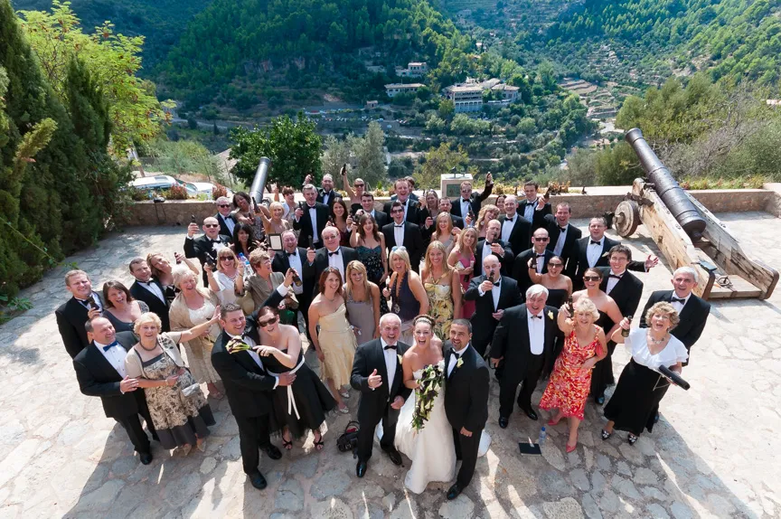 Large wedding group posing outdoors on a stone terrace, with the bride and groom in front, surrounded by guests in formal attire and two historical cannons on either side, overlooking a green mountainous landscape.