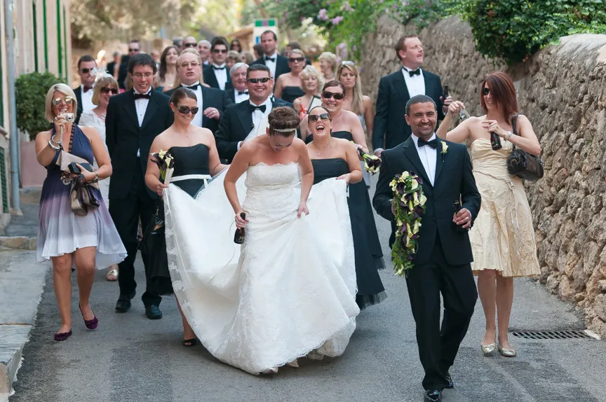Bride in white gown and groom in black tuxedo walking down a street with wedding party and guests behind them, all smiling and celebrating.