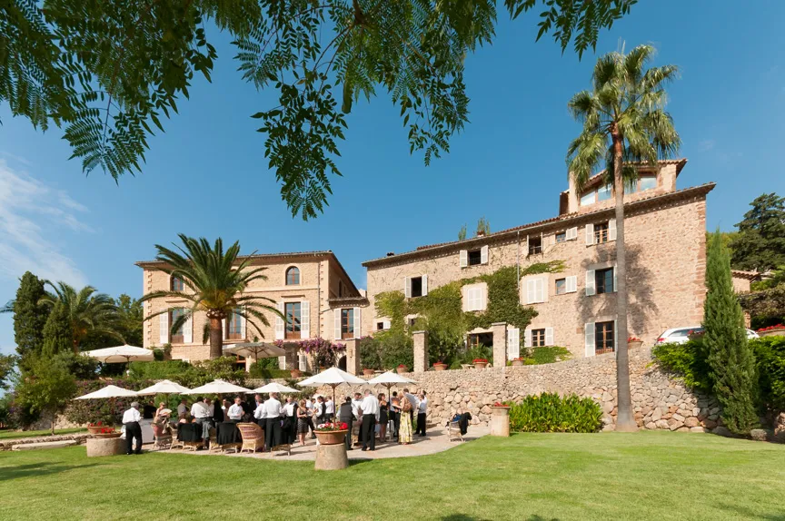 Outdoor gathering of people under white umbrellas on a patio in front of a large stone building with palm trees and clear blue sky.
