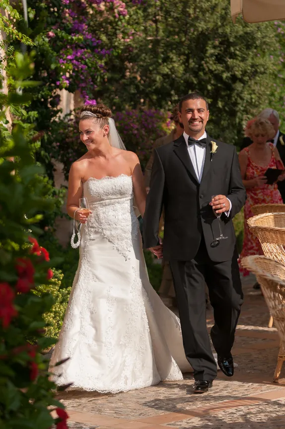Bride in a white lace wedding dress and groom in a black tuxedo holding hands and smiling outdoors with guests in the background.