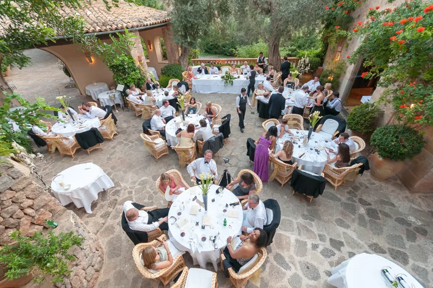 Outdoor wedding reception with guests seated at round tables covered in white linens on stone patio surrounded by greenery and flowers.