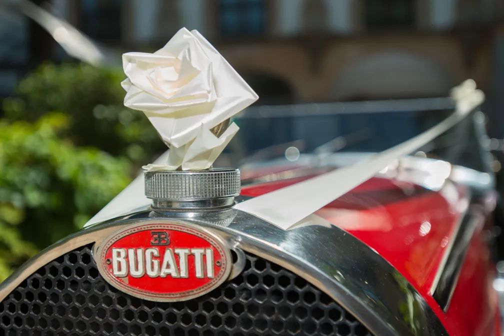 Close-up of a red Bugatti car grille with a white paper flower attached to the hood ornament.