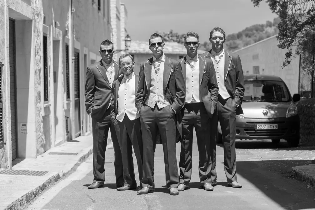 Five men in tuxedos and sunglasses standing together on a sunlit street with a car in the background.