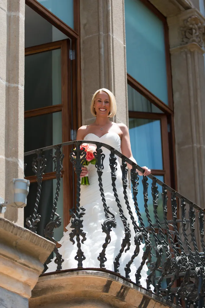 Bride in a strapless white wedding gown holding a bouquet of orange and pink flowers, standing on a wrought iron balcony in front of large windows.