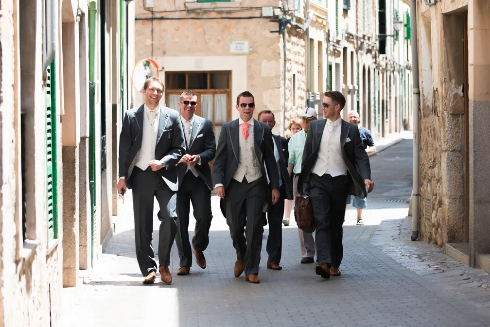 Four men in formal suits walking and smiling down a sunny narrow cobblestone street with other casually dressed pedestrians behind them.