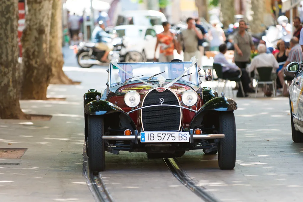 Vintage red and black Bugatti convertible car driving on a street with tram tracks and blurred pedestrians in the background.