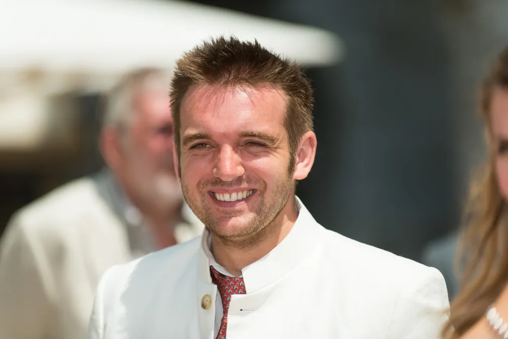 Smiling man with short spiked brown hair wearing a white jacket and red patterned tie in a sunny outdoor setting.