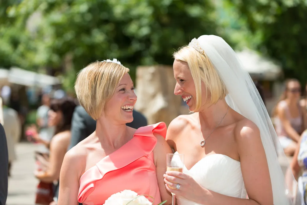 Bride in white dress and veil smiling and holding a glass, laughing with a woman in a coral one-shoulder dress holding a bouquet.