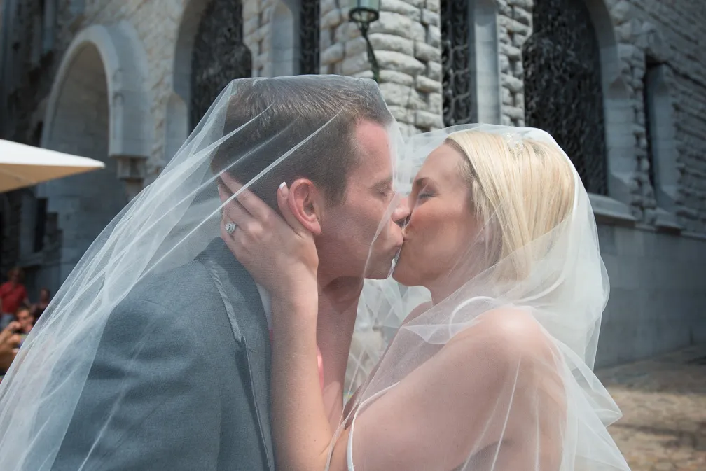 Bride and groom kissing under a wedding veil outside a stone building with arched windows.