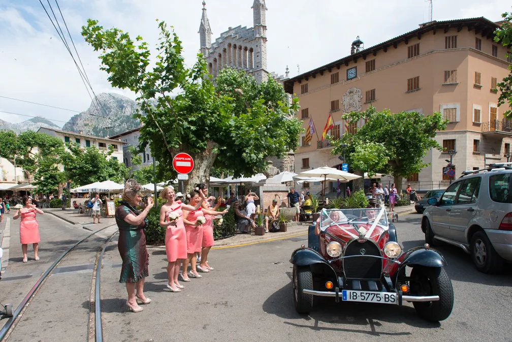 Vintage convertible car driving past a group of bridesmaids in coral dresses and a woman in a teal dress on a sunny street with trees and historic buildings.