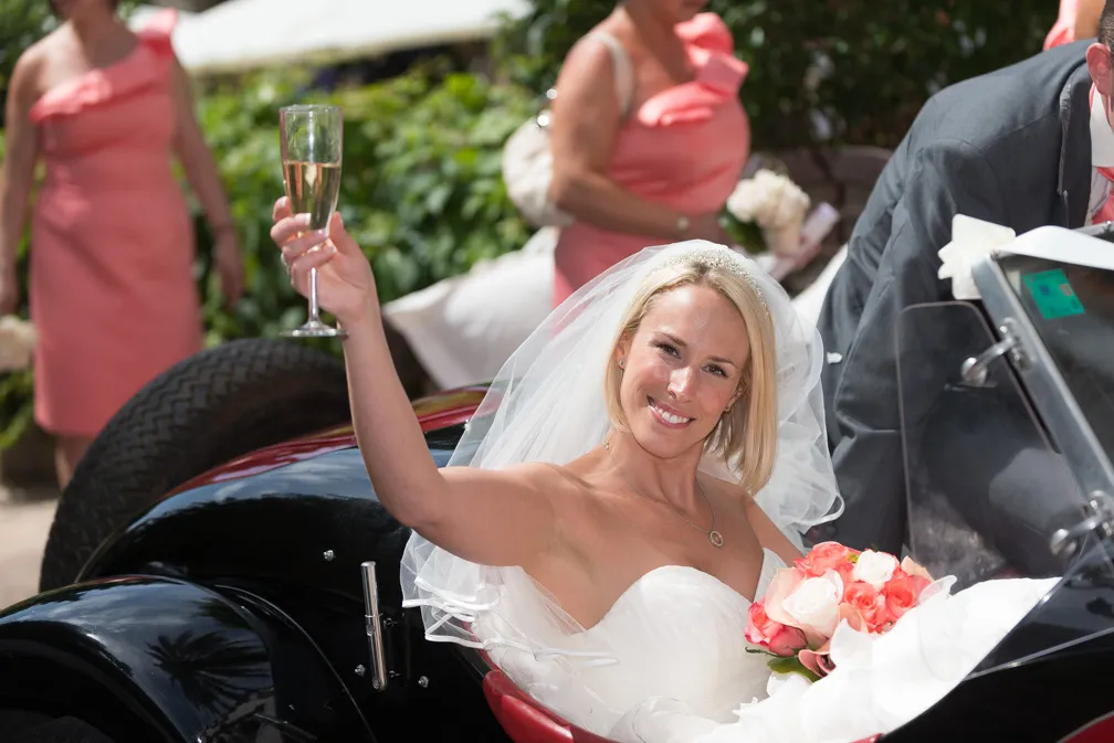 Smiling bride in white dress and veil raising a champagne glass while sitting in a vintage car, holding a bouquet of pink and white roses.