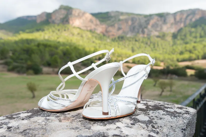 Pair of elegant white high-heeled sandals with rhinestone straps placed on a stone surface with a mountainous landscape in the background.