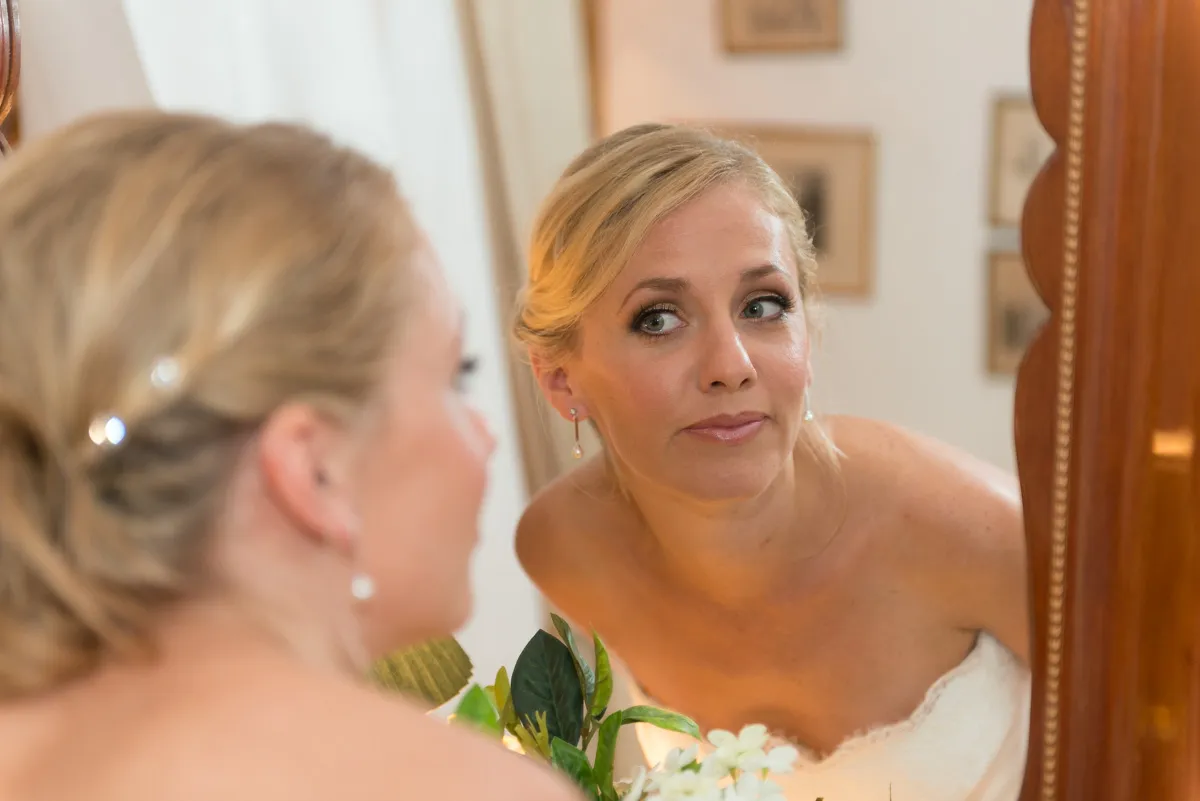 Bride with blonde hair and pearl earrings looking at herself in a wooden framed mirror while holding a bouquet of white flowers and green leaves.