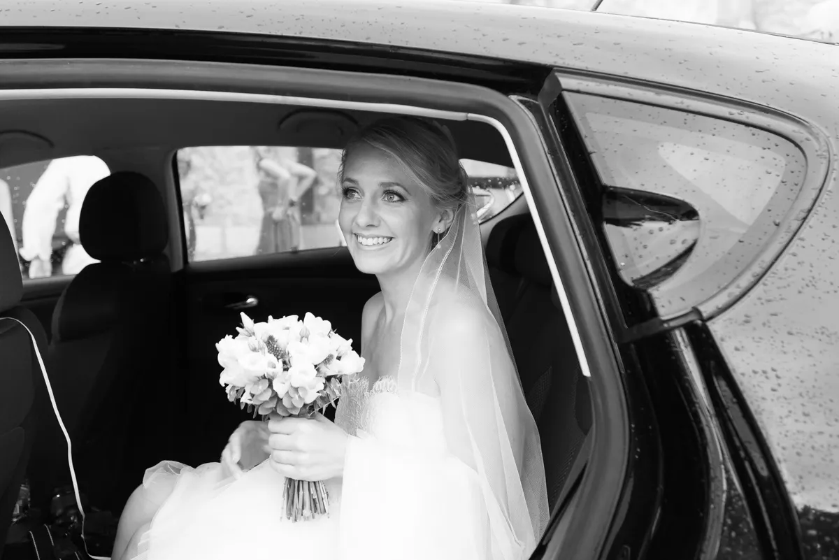 Smiling bride in a wedding dress and veil holding a bouquet, sitting in the backseat of a car with rain droplets on the window.