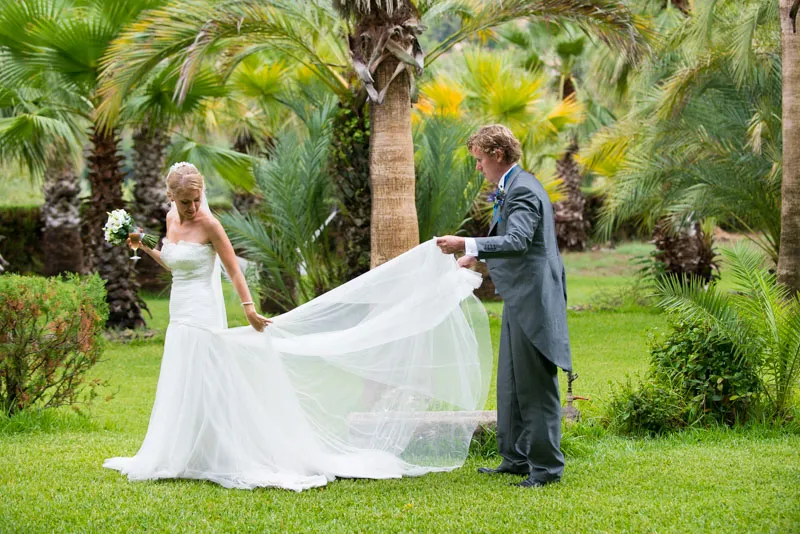 Bride in a white wedding dress holding a bouquet, with groom in a gray suit adjusting her dress in a lush green garden with palm trees.