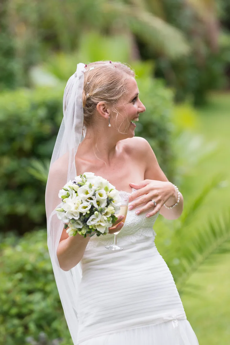 Smiling bride in a strapless white dress holding a bouquet of white flowers and a glass of wine outdoors.