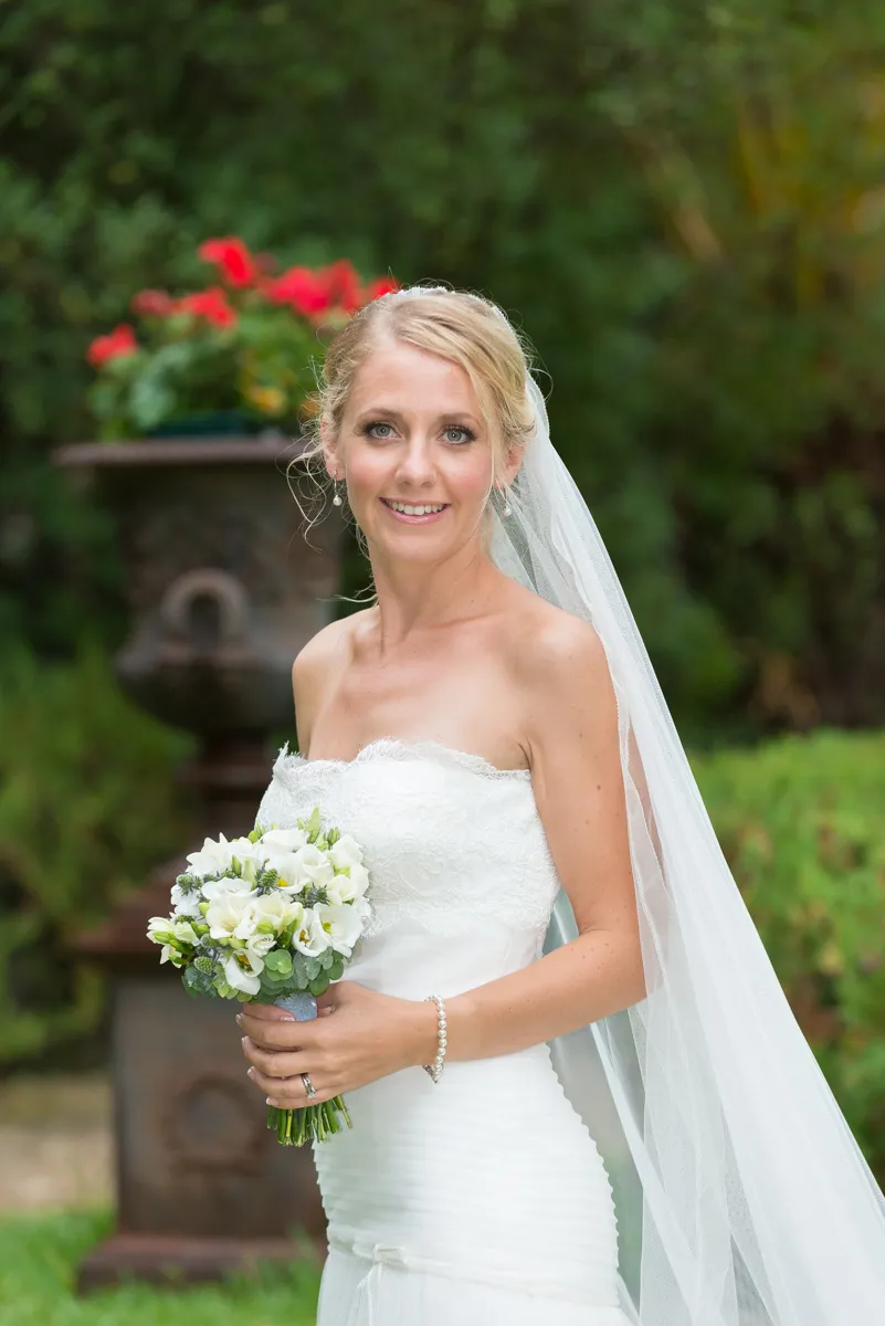 Bride in a strapless white wedding dress holding a bouquet of white flowers, standing outdoors with greenery in the background.