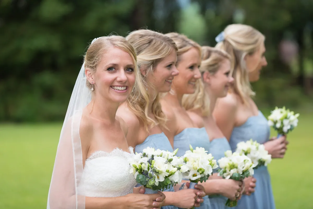 Bride in a white strapless dress and veil smiling with four bridesmaids in blue dresses holding white flower bouquets in a garden.