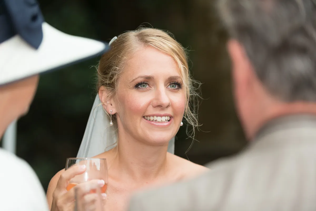 Smiling blonde bride holding a glass and talking to two people at her wedding.