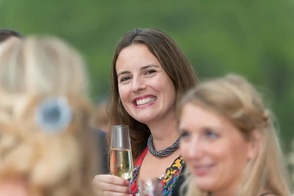 Smiling woman holding a glass of champagne at an outdoor social event with blurred people in the foreground.