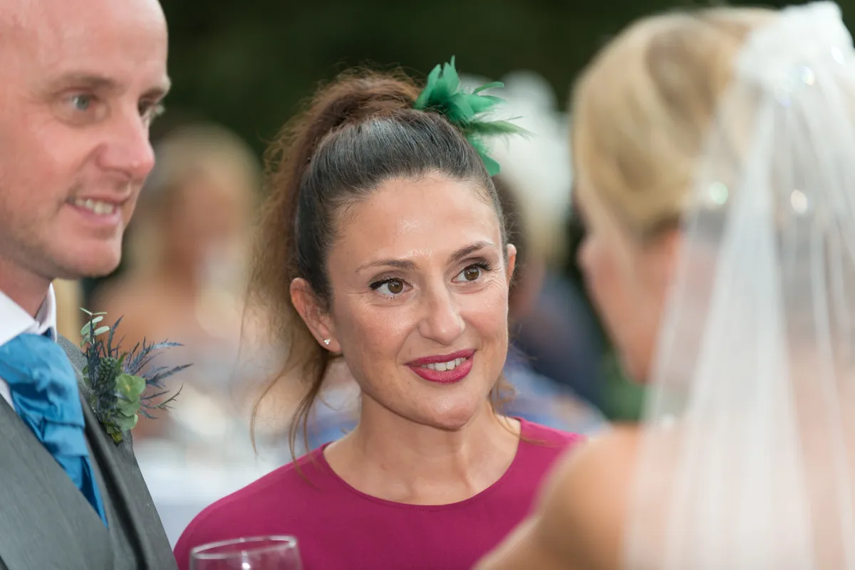A smiling woman in a purple dress with a green hair accessory, talking to a man in a suit with a blue tie and a person in a wedding veil.